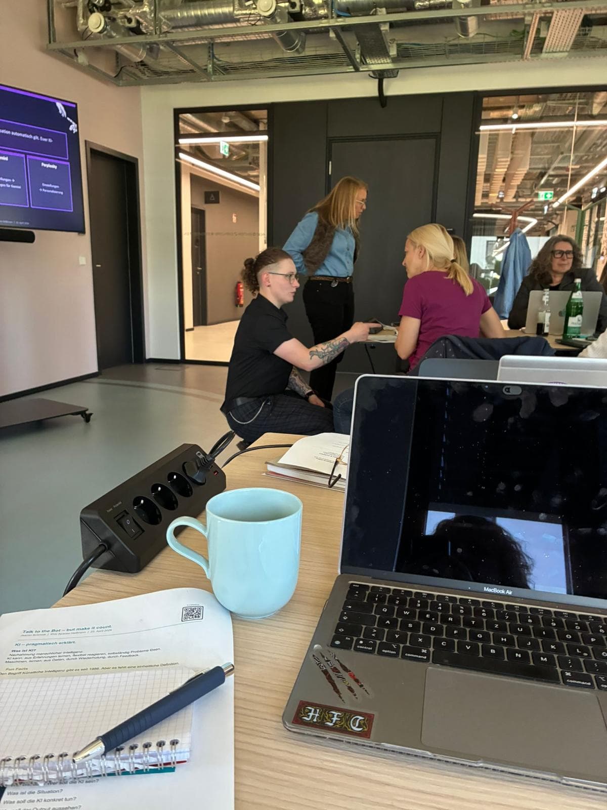 Workshopsituation im Alan-Turing-Raum: Jasmin Schmidt im Gespräch mit Teilnehmerinnen an den Arbeitstischen, im Hintergrund eine Präsentationsfolie
